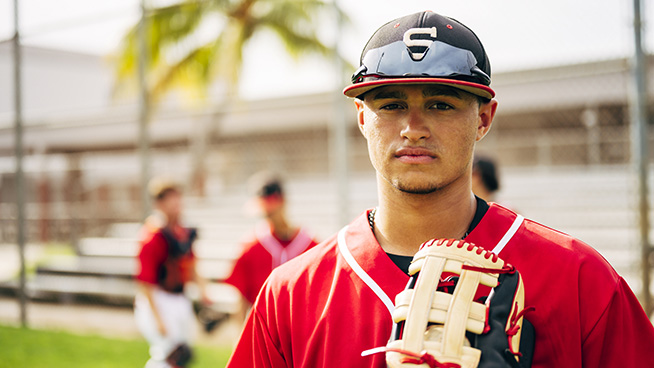 Close-up of young and serious Hispanic baseball player with glove standing outdoors and waiting for the game to begin.