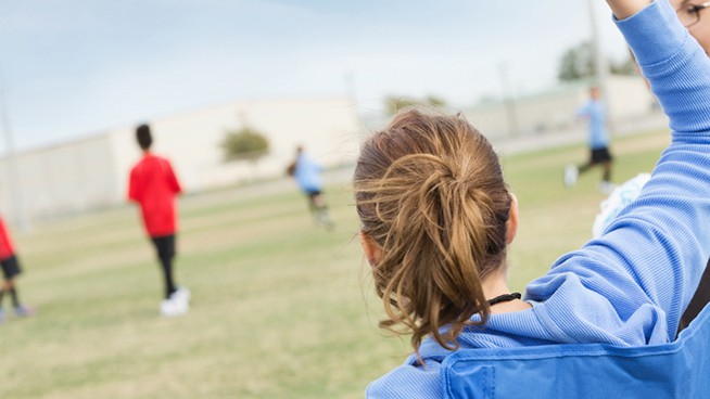 Soccer mom waving pompoms, cheering for kids during game.