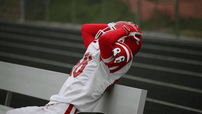 young football athlete showing frustration by grabbing helmet while sitting on bench