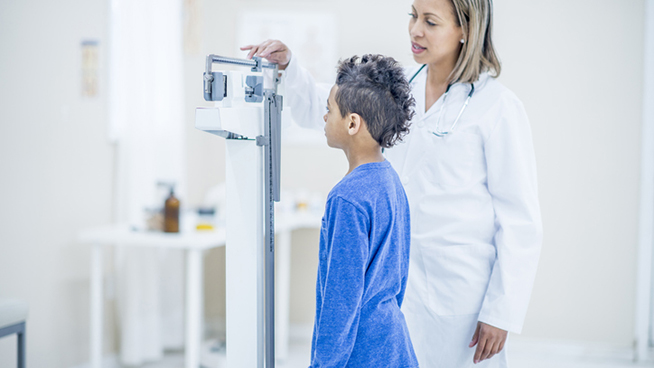 A female doctor and young boy are indoors in a medical office. The doctor is taking the boy's measurements while he stands on a scale.