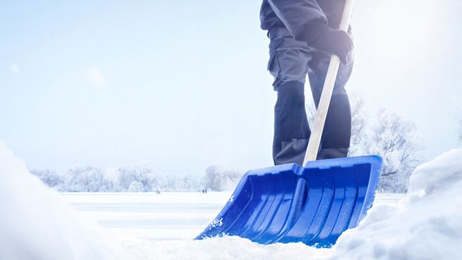A person using a snow shovel in a snowy landscape. Only the lower part of the person is visible. Low angle shot with snow piles in the foreground.