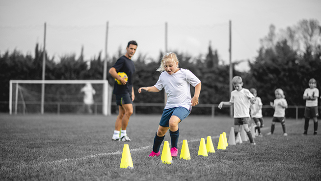 Focused 7 year old girl moving around row of pylons as she and teammates do agility drill under direction of male coach during sports training camp.