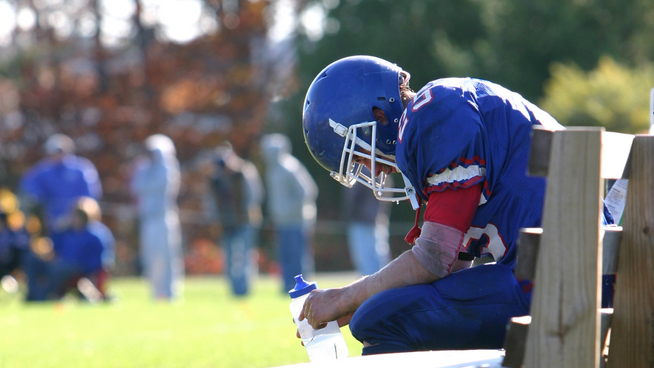 football athlete sitting on bench with head down and holding water bottle at outdoor football game