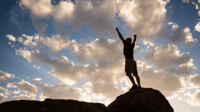 male standing on top of large rock in the wilderness with hands raised above his head in confidence