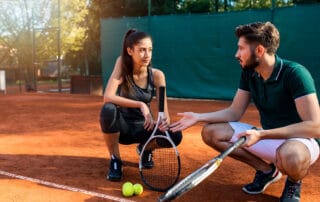 male tennis coach squatting down on court with female tennis athlete while coaching