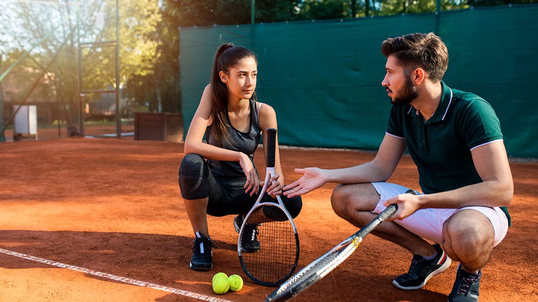 male tennis coach squatting down on court with female tennis athlete while coaching